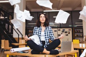 A young woman is sitting on a desk in a meditation pose with her eyes closed as papers fly about around her