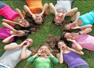 A group of young people laying on their backs in a circle with all their heads facing towards the centre of the circle, cupping their mouths with their hands and shouting