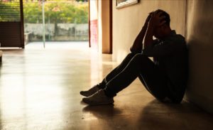 A young man is sitting on the floor with his back against a wall with his head in his hands