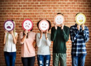 Young people against a brick wall with cartoon paper plate faces featuring different emotions covering their own faces