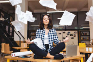 teenage girl sitting cross-legged on a school desk in a classroom meditating with papers flying all around her