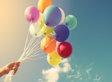 A photo of blue sky and white clouds, and a big bunch of multi-coloured balloons flying in the air being held together with a bundle of string that is being held by a hand coming up from the bottom left corner of the image.