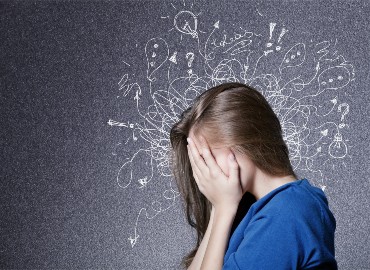 A photo of a teenage girl facing sideways with her hands covering her face, and on the background all around her head are chalk drawn squiggly lines in all directions, some with words such as 'ideas', lightbulbs, exclamation marks, question marks, arrows, ellipses.