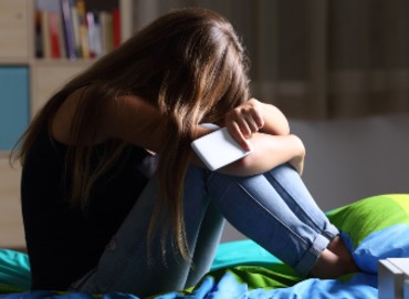 A girl in her room with her legs tucked up and hiding her face. She is holding her mobile phone