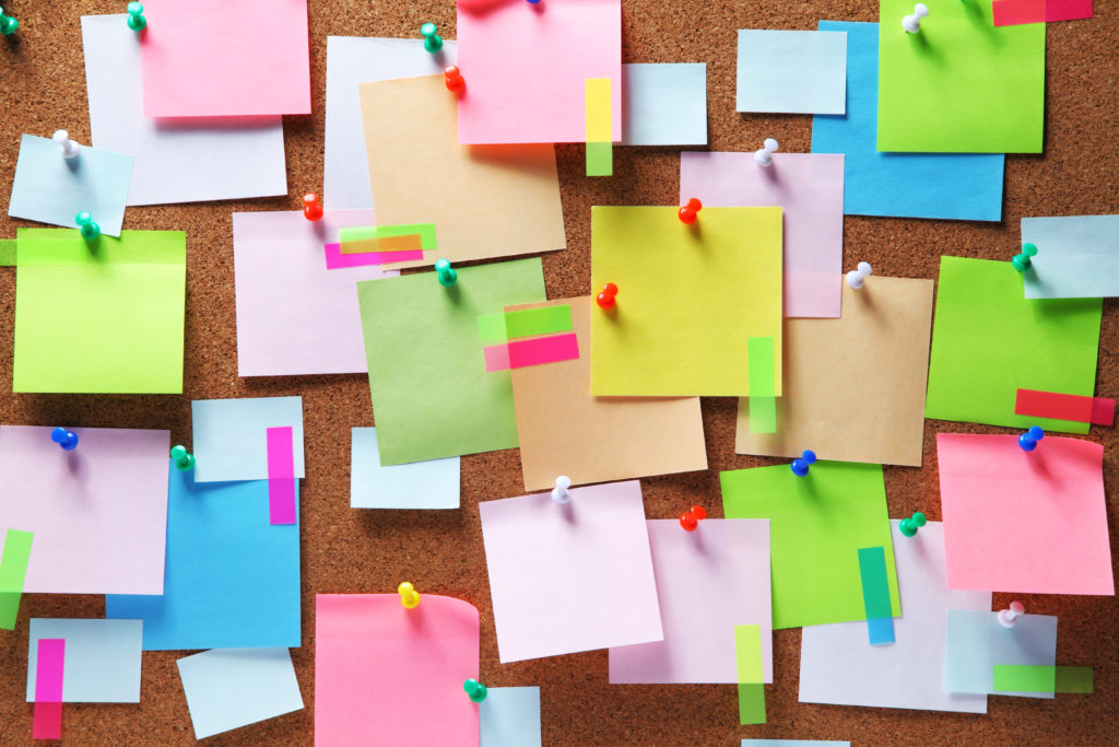 A brown cork-board background with a variety of colourful post-it notes stuck to the board with colourful pins